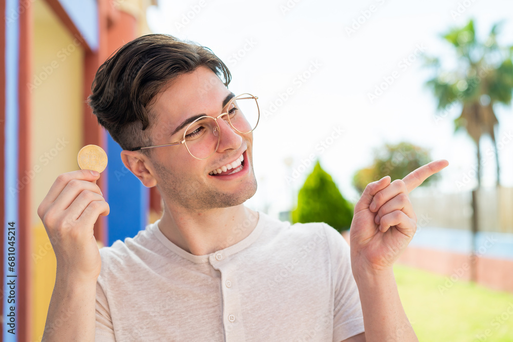 Young handsome man holding a Bitcoin at outdoors pointing to the side to present a product Young handsome man holding a Bitcoin at outdoors pointing to the side to present a product