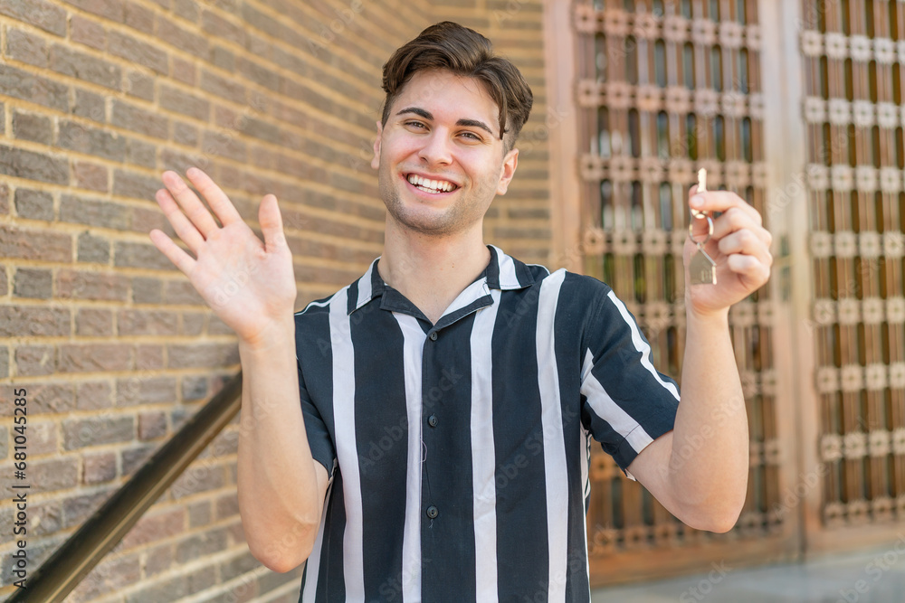 Naklejka premium Young handsome man holding home keys at outdoors saluting with hand with happy expression