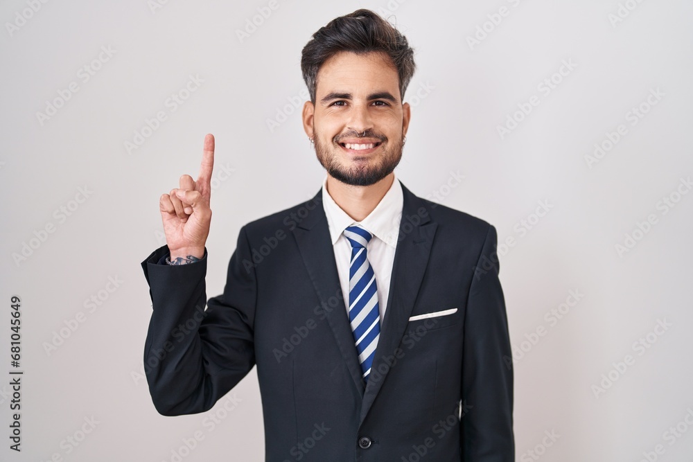 Young hispanic man with tattoos wearing business suit and tie showing and pointing up with finger number one while smiling confident and happy. Young hispanic man with tattoos wearing business suit and tie showing and pointing up with finger number one while smiling confident and happy.