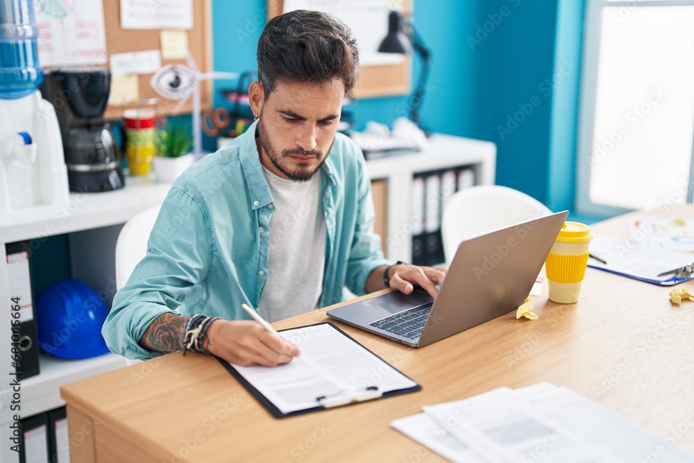 Young hispanic man business worker using laptop writing on document at office