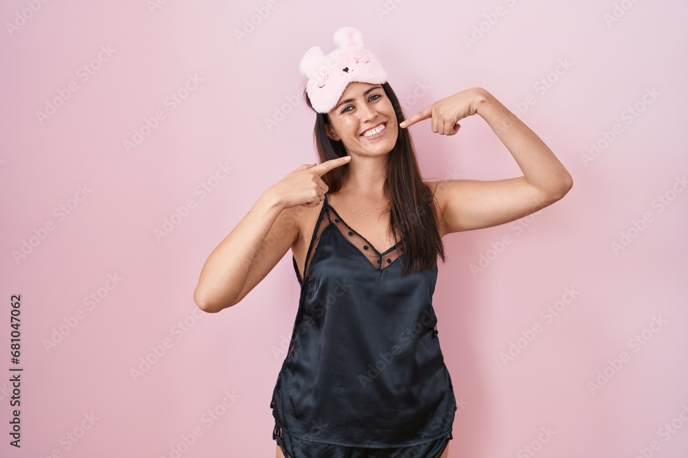 Young brunette woman wearing sleep mask and pyjama smiling cheerful showing and pointing with fingers teeth and mouth. dental health concept. Young brunette woman wearing sleep mask and pyjama smiling cheerful showing and pointing with fingers teeth and mouth. dental health concept.