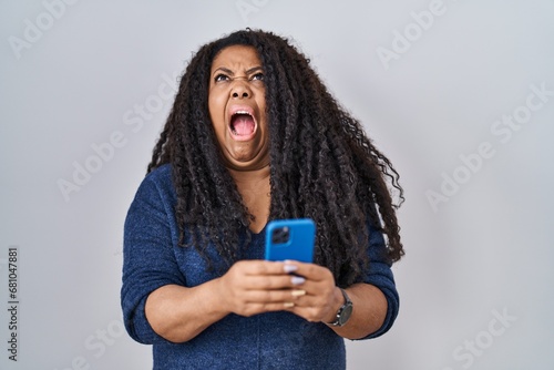 Plus size hispanic woman using smartphone typing message angry and mad screaming frustrated and furious, shouting with anger looking up. photo