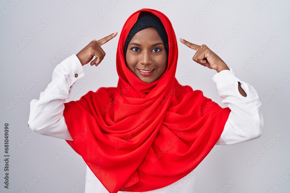 Young arab woman wearing traditional islamic hijab scarf smiling pointing to head with both hands finger, great idea or thought, good memory Young arab woman wearing traditional islamic hijab scarf smiling pointing to head with both hands finger, great idea or thought, good memory