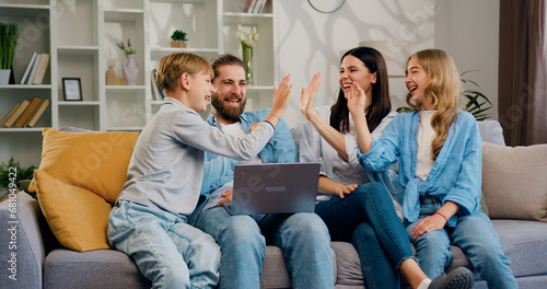 Fototapeta Naklejka Na Ścianę i Meble -  Happy overjoyed attractive young family siting on sofa check email on laptop celebrate amazing news, giving high five, congratulate each other with success. Men, women and children have good mood