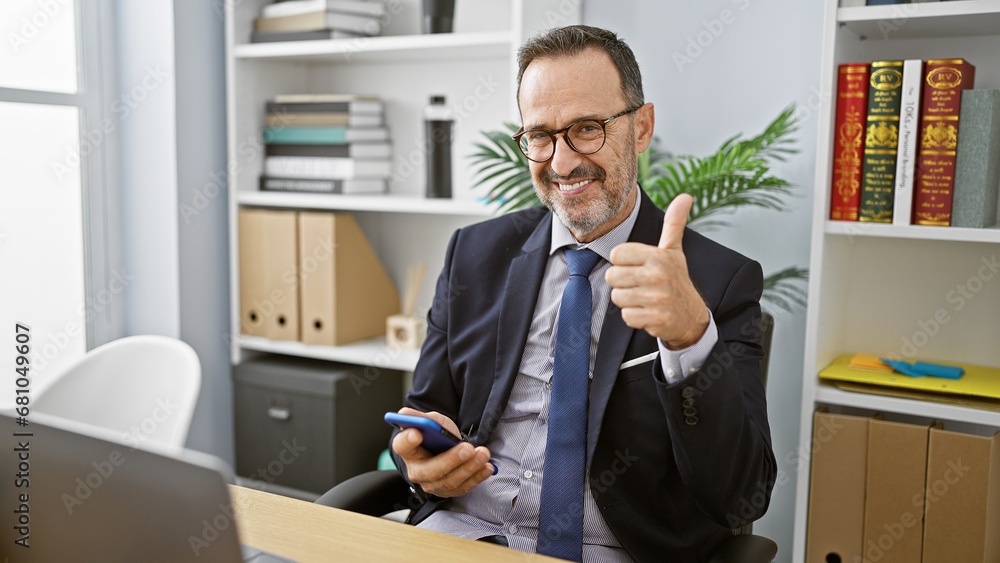 Cheerful, grey-haired middle age man, a diligent business worker, works his smartphone, giving an assertive thumb up gesture in his office room, projecting a confident, positive aura! Cheerful, grey-haired middle age man, a diligent business worker, works his smartphone, giving an assertive thumb up gesture in his office room, projecting a confident, positive aura!
