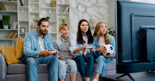 Happy smiling family of mother, father, son, and daughter playing video game together sitting on a couch at home. Parents play online using a controller and children cheer. Relationship, bonding