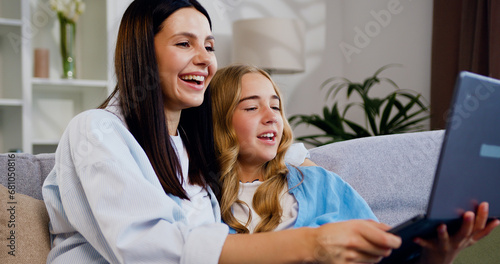 Portrait of happy woman using laptop computer, showing video content, explaining online app work, teaching cute little daughter. Family love and happy mother and girl with computer on couch for