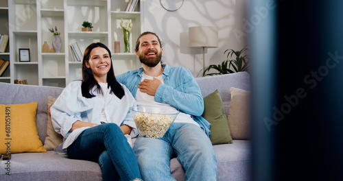 Portrait of young couple spending time at home, sitting on sofa watching exciting TV show eating popcorn. Happy young love marriage, woman leisure time snack on couch and man smiling watch streaming