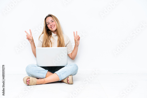 Young woman with laptop sitting on the floor isolated on white background showing victory sign with both hands © luismolinero