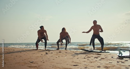 A bare-chested man in a wetsuit shows his friends how to surf properly on the Sandy Beach near the sea in summer. Two guys are learning how to swim with a surf and their friend is helping them with photo