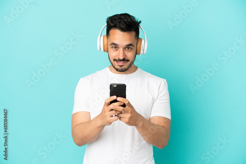 Young caucasian man isolated on blue background listening music with a mobile and looking front