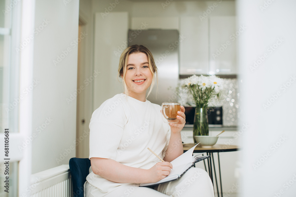 Smiling young woman drinking tea and writing in her diary or creative journal in the morning in the kitchen. Smiling young woman drinking tea and writing in her diary or creative journal in the morning in the kitchen.