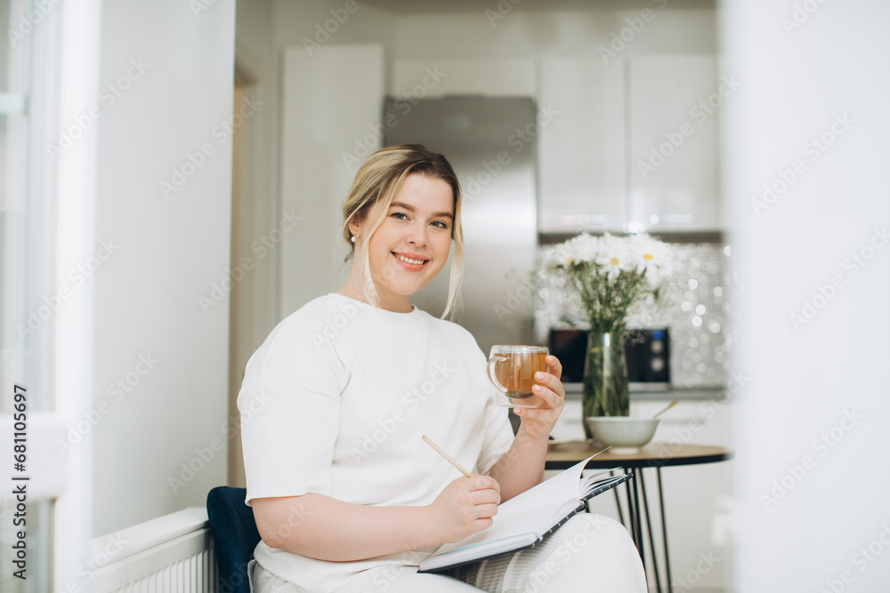 Smiling young woman drinking tea and writing in her diary or creative journal in the morning in the kitchen. Smiling young woman drinking tea and writing in her diary or creative journal in the morning in the kitchen.