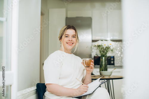 Smiling young woman drinking tea and writing in her diary or creative journal in the morning in the kitchen.