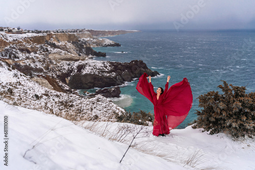 Woman red dress snow sea. Happy woman in a red dress in the snowy mountains by the emerald sea. The wind blows her clothes, posing against sea and snow background.