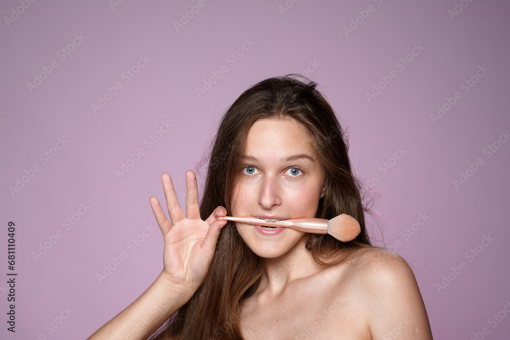 Woman holding a makeup brush in her mouth. Seminude woman in studio on pink background in pink neon light. Concept of beauty, cosmetology, skin care. Woman holding a makeup brush in her mouth. Seminude woman in studio on pink background in pink neon light. Concept of beauty, cosmetology, skin care.