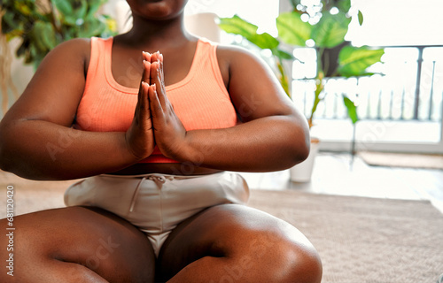 Sport for relaxation. Close up of overweight african woman keeping hands in namaste gesture and sitting on lotus pose while meditating at home, copy space. Concept of yoga, wellbeing and spirituality.
