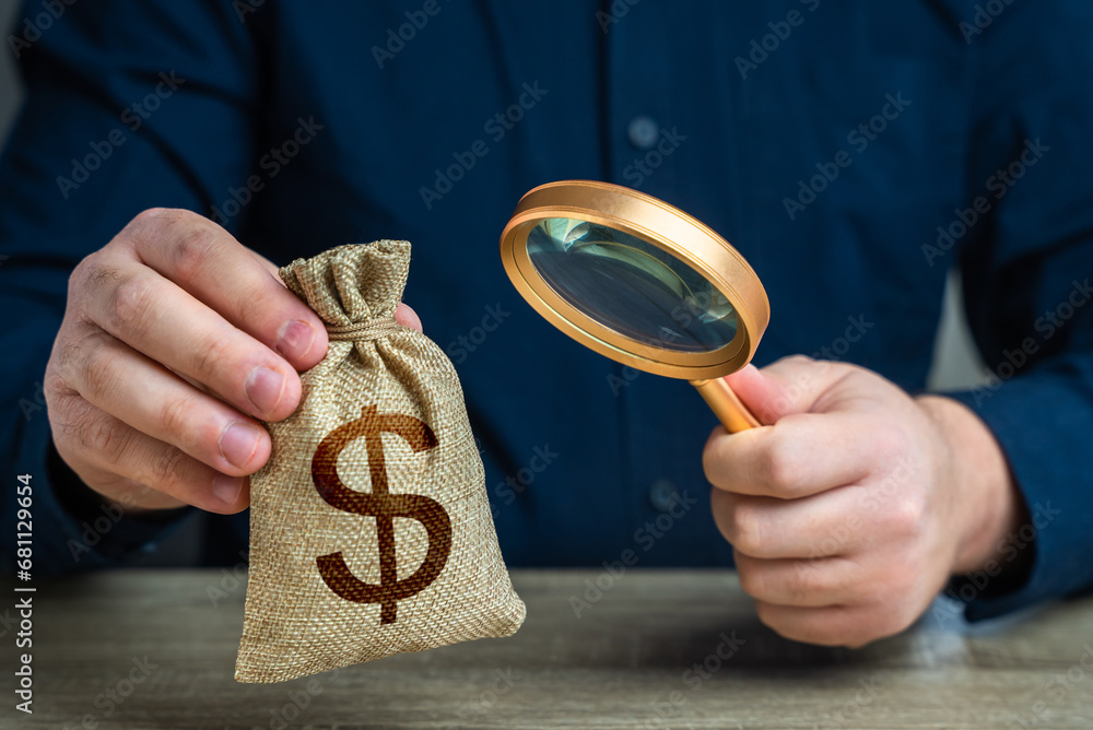 A man examines a dollar money bag through a magnifying glass. Investigating capital origins. Anti money laundering, tax evasion. Study terms and conditions on deposit or loan. Find investments - obrazy, fototapety, plakaty 