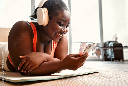 Motivation in sport. Black lady lying on stomach on yoga mat and listening music in wireless headphones. Smiling plump female in active outfit using cell phone with favorite playlist during training. photo