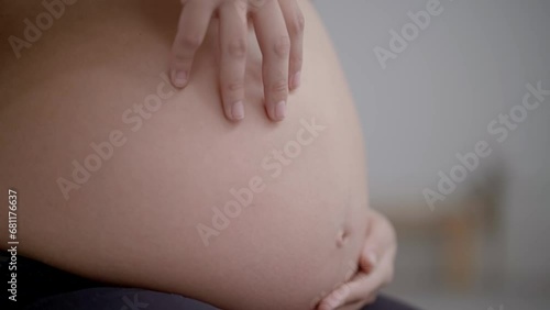Unrecognizable pregnant Caucasian woman in sportswear doing yoga pose indoors. White female touching her huge belly. Fitness people expecting a baby. Unfocused background.