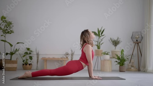 Full body side view of a young blonde woman in a red sports outfit exercising in a modern yoga or pilates centre or at home. Doing pose heathy living and self-care.