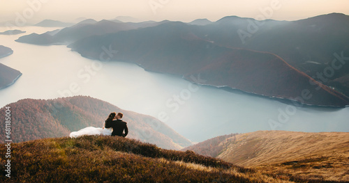 A young wedding couple is sitting on a mountain, with a mountain and a bay in the background. The couple hugs and looks at the landscape. Autumn weather. A wonderful view