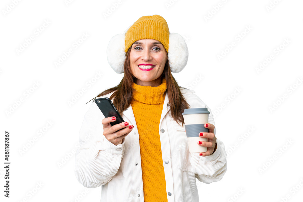 Middle age woman wearing winter muffs over isolated chroma key background holding coffee to take away and a mobile Middle age woman wearing winter muffs over isolated chroma key background holding coffee to take away and a mobile