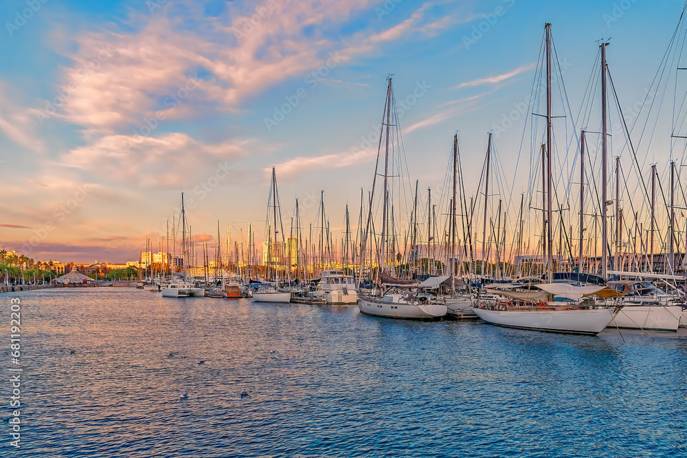 Yachts moored in the Port of Barcelona at sunset, Spain. Many boats with masts in the bay of the Mediterranean Sea against the backdrop of the city coastline illuminated by the rays of the setting sun - obrazy, fototapety, plakaty 