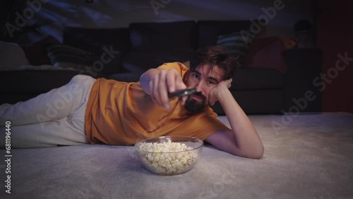 Young European man with beard lying on living room carpet eating popcorn as he changing channel smiling happily. Millennial guy enjoying movie or series and zapping with bowl inside home at night