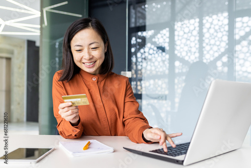 Portrait of young successful satisfied Asian woman inside office at workplace, business woman smiling, using bank credit debit card for online shopping, working with laptop.
