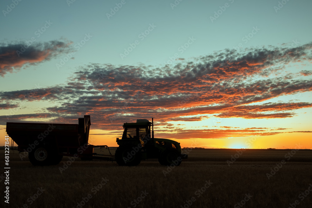 Tractor and grain cart parked in a backlit field as the sun sets. Sky with clouds in orange tones. Copy space Tractor and grain cart parked in a backlit field as the sun sets. Sky with clouds in orange tones. Copy space