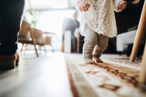 A baby's tiny bare feet take careful steps on a patterned rug, guided by an adult's hands, in a warm, sunlit room.