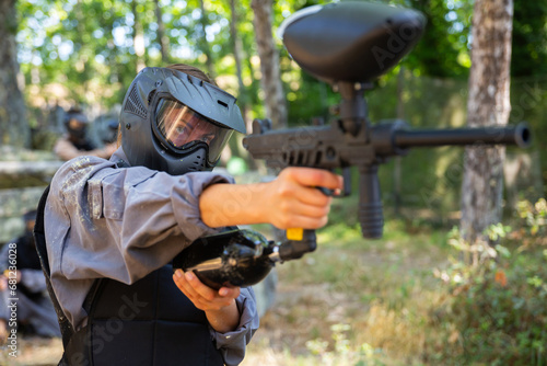 Woman in protective clothes and helmet aiming with paintball marker during team fight on battleground.