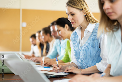 Portrait of concentrated womens and mens studying in classroom  listening to teacher and writing in notebook
