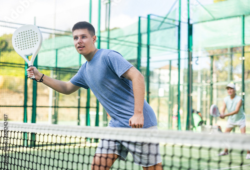 Young man in shorts and t-shirt playing padel tennis on court. Racket sport training outdoors. © JackF