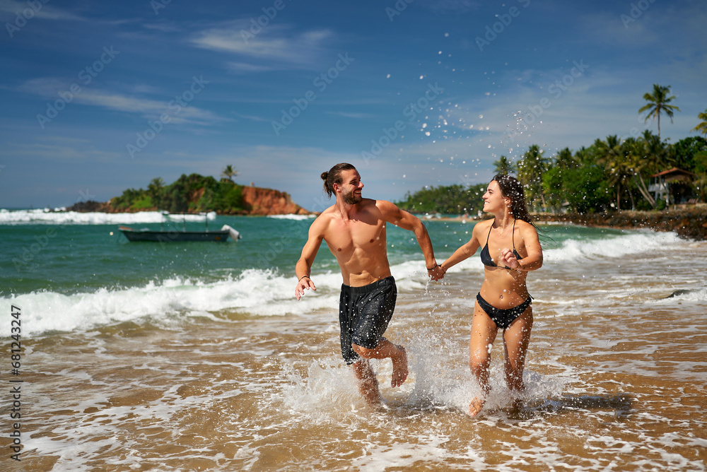 Playful couple runs on tropical beach, hand in hand, splashing water, joyous moments together. Fit man, slender woman enjoy sunny seaside, engaged in active lifestyle, vacation vibes, fitness.