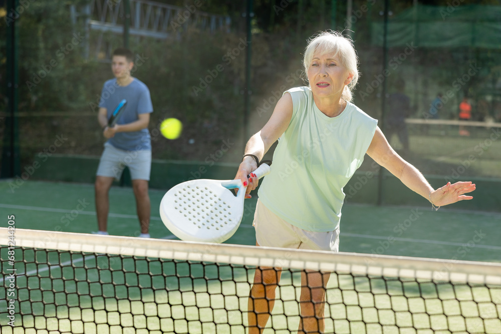 Focused aged woman playing friendly paddleball match on outdoor summer court. Senior people sports concept..