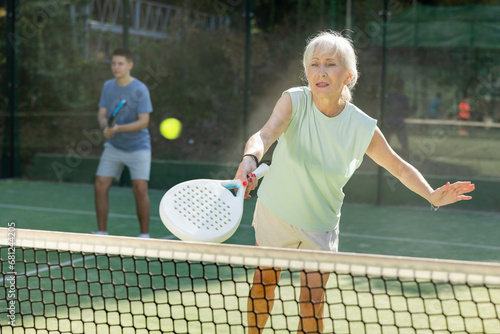 Focused aged woman playing friendly paddleball match on outdoor summer court. Senior people sports concept.. © JackF