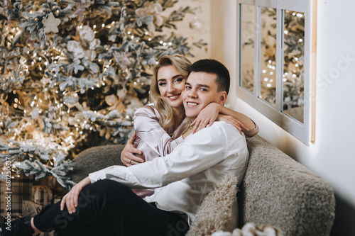 A stylish young man and a beautiful smiling woman are hugging while sitting on a sofa in a room with a festive decorated interior, decor for Christmas, New Year. Photography, portrait. © shchus