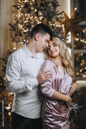 A young man and a beautiful happy smiling woman in a dress are hugging in a room with a festive decorated interior, decor for Christmas, New Year on the background of a Christmas tree with a garland.