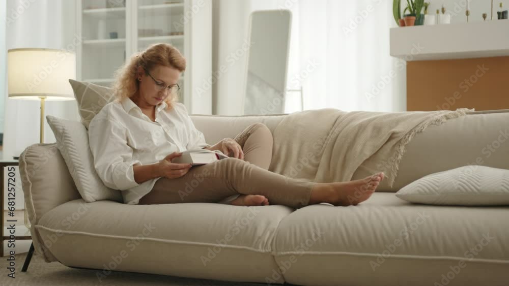 Tired lady in glasses with thick book in hands sitting with streched legs on sofa, closing eyes, benting head over the book. Light room with renovation. High quality 4k footage Tired lady in glasses with thick book in hands sitting with streched legs on sofa, closing eyes, benting head over the book. Light room with renovation. High quality 4k footage