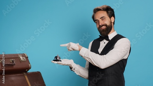 Hotel porter points to service bell, advertising concierge assistance and ringing to provide luxury service with baggage. Young professional doorman helping with trolley bags, hospitality industry. photo