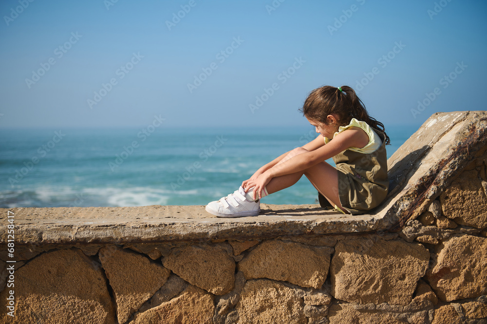 Full size portrait of a cute little kid girl sitting on a stone parapet and dreamily looking aside admiring Ocean storm Full size portrait of a cute little kid girl sitting on a stone parapet and dreamily looking aside admiring Ocean storm