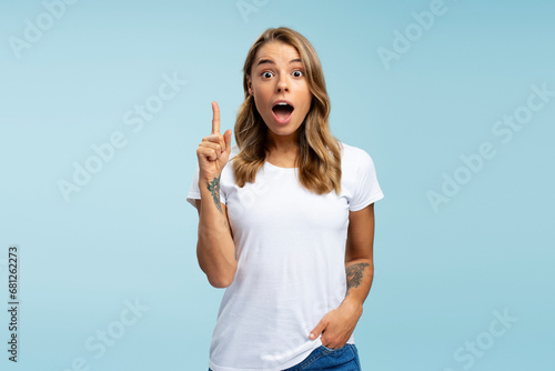 Funny emotional student holding finger up having creative idea isolated on blue background, education concept. Amazed attractive young woman with open mouth pointing hand looking at camera in studio photo