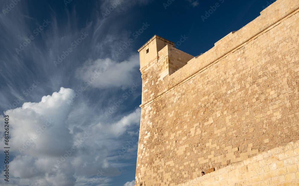 Close-up of the high wall of the historic citadel on Gozo in Malta. The strong walls tower high up. The sky is blue with light-coloured clouds. Close-up of the high wall of the historic citadel on Gozo in Malta. The strong walls tower high up. The sky is blue with light-coloured clouds.
