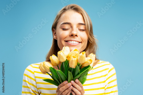 Closeup portrait of cute smiling woman with eyes closed holding bouquet of beautiful yellow tulips flowers isolated on blue background. Natural beauty, spring time, birthday gift concept