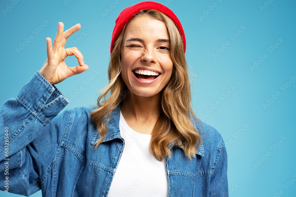Smiling beautiful woman winking wearing stylish red hat showing ok  sign isolated on blue background. Portrait happy modern hipster female with white teeth looking at camera in studio, dental concept  - obrazy, fototapety, plakaty 