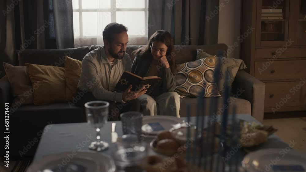 Full shot of bearded Jewish man in kippah sitting on couch with young girl, holding Torah book, reading and discussing holy scripture, and festive table with hanukkiah in blurred foreground Full shot of bearded Jewish man in kippah sitting on couch with young girl, holding Torah book, reading and discussing holy scripture, and festive table with hanukkiah in blurred foreground