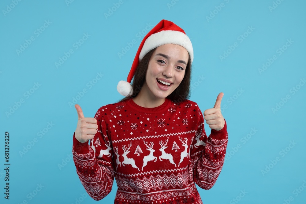 Happy young woman in Christmas sweater and Santa hat showing thumbs up on light blue background Happy young woman in Christmas sweater and Santa hat showing thumbs up on light blue background