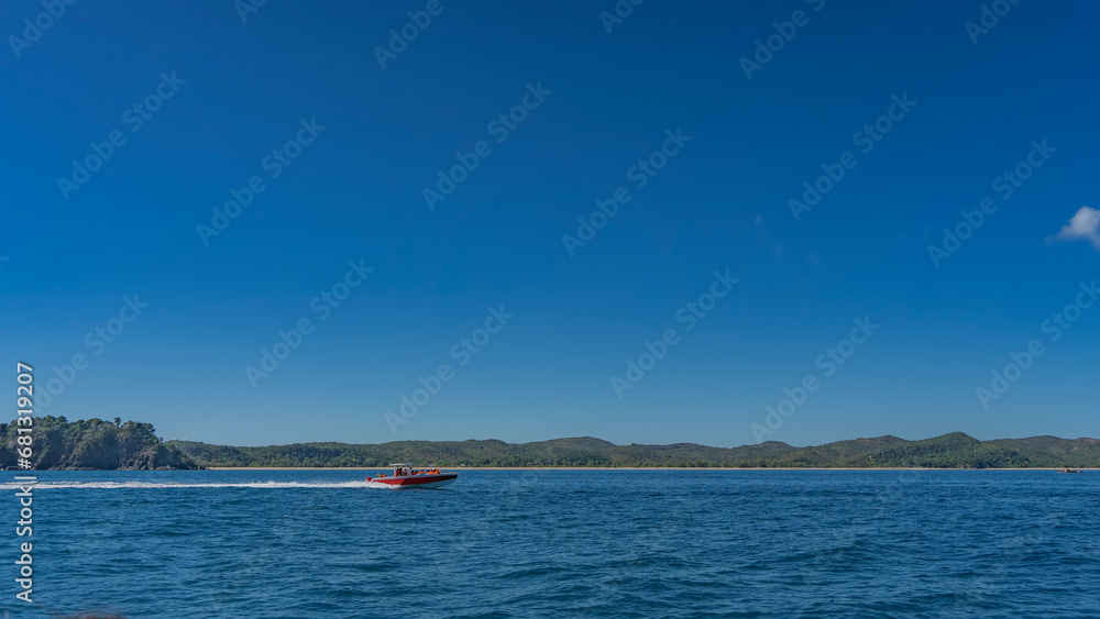 A boat with tourists is rushing across the blue ocean. Foam trail behind the stern. Green coastal hills against the azure clear sky in the distance. copy space. Madagascar.